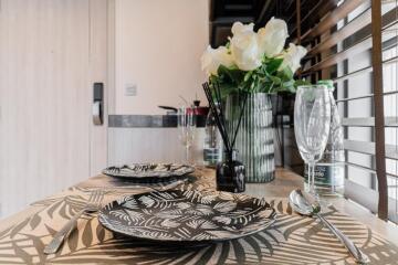 Table set for two with plates, glasses, and cutlery, next to a window with blinds and a vase of white flowers