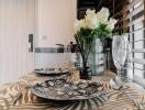 Table set for two with plates, glasses, and cutlery, next to a window with blinds and a vase of white flowers