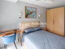 Bedroom with modern wooden wardrobe and bed, desk, blue chair, painting on the wall, and overhead lighting