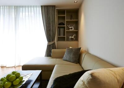 A cozy living room with a sectional sofa and a coffee table adorned with green apples, large window with curtains, and a small shelving unit.