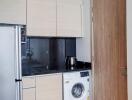 Compact kitchen with wooden cabinetry, stainless steel refrigerator, washing machine, and wood-paneled door