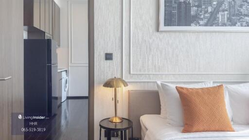 Modern bedroom with white walls, bed, and orange pillow adjacent to a kitchen area