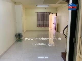 An unfurnished living room with tiled flooring, a wall-mounted fan, curtained window, and staircase leading to another floor.