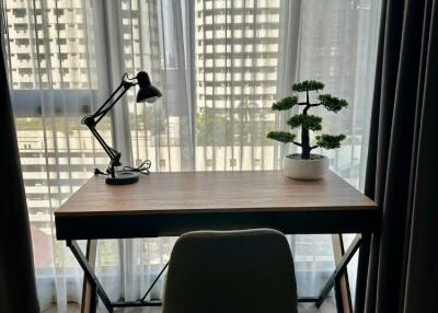 A study area with a desk, chair, lamp, and plant in front of a large window with city view.