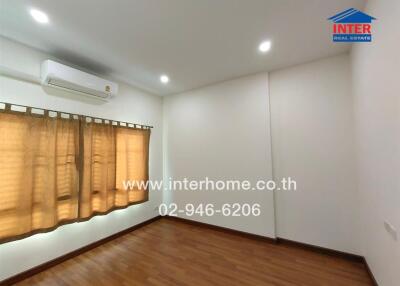 A well-lit empty bedroom with wooden floors, an air conditioning unit, and beige curtains covering the window.