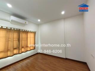 A well-lit empty bedroom with wooden floors, an air conditioning unit, and beige curtains covering the window.