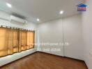 A well-lit empty bedroom with wooden floors, an air conditioning unit, and beige curtains covering the window.