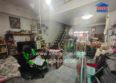 Cluttered living room with a gaming chair, stairs, books, and various personal items.