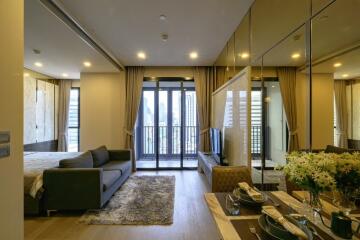 Modern apartment living room with a view of the city, featuring a couch, dining table, and large windows leading to a balcony.