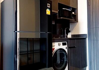 Modern kitchen area with black cabinets, refrigerator, washing machine, microwave, and sink