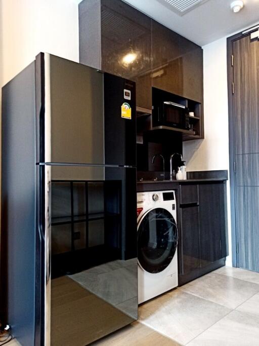 Modern kitchen area with black cabinets, refrigerator, washing machine, microwave, and sink