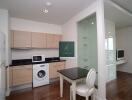 Modern kitchen with light wood cabinetry, a washing machine, microwave, refrigerator, and a dining area with a table and a chair.