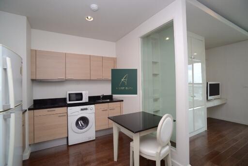 Modern kitchen with light wood cabinetry, a washing machine, microwave, refrigerator, and a dining area with a table and a chair.