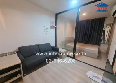 Living area with adjacent bedroom separated by glass partition, featuring a sofa, side table, and air conditioning.