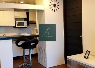 Modern kitchen and living area with bar stools, wall clock, and flat-screen TV