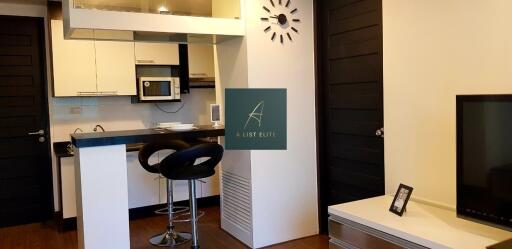 Modern kitchen and living area with bar stools, wall clock, and flat-screen TV