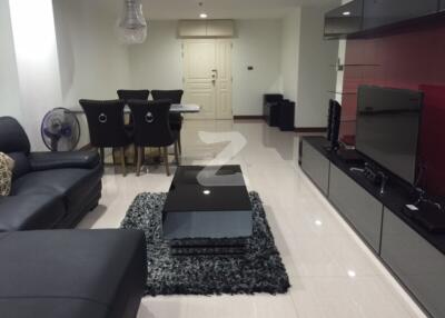 Modern living room with black leather sectional sofa, glass coffee table, flat-screen TV, and dining area with upholstered chairs.