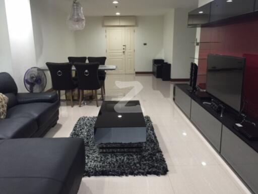 Modern living room with black leather sectional sofa, glass coffee table, flat-screen TV, and dining area with upholstered chairs.