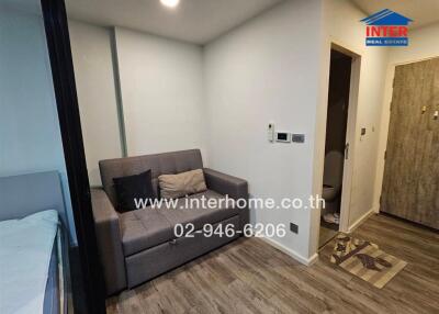 Small apartment living area with sofa, partial view of bed, and hallway with wooden flooring