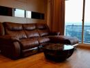 Living room with brown leather sectional sofa, glass-top coffee table, and a large window with a view.