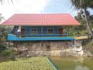 Front view of a colorful house with a red roof, elevated foundation, and a nearby pond