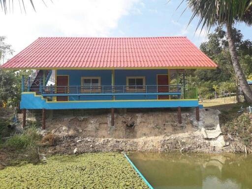 Front view of a colorful house with a red roof, elevated foundation, and a nearby pond