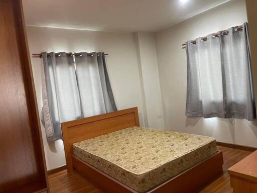 A cozy bedroom with a wooden bed frame and mattress, two windows covered with gray curtains, and a wooden wardrobe.