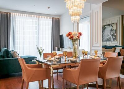 A modern living room and dining area with a glass dining table, leather chairs, a chandelier, and a large window with curtains.