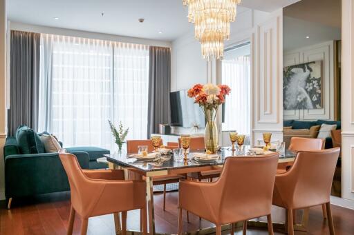 A modern living room and dining area with a glass dining table, leather chairs, a chandelier, and a large window with curtains.