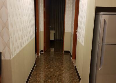 A well-lit hallway with tiled flooring and a refrigerator.