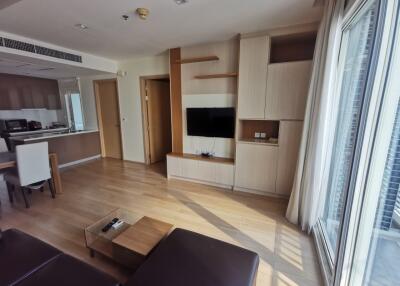 Modern living room with TV and wooden furniture, adjacent to an open kitchen.