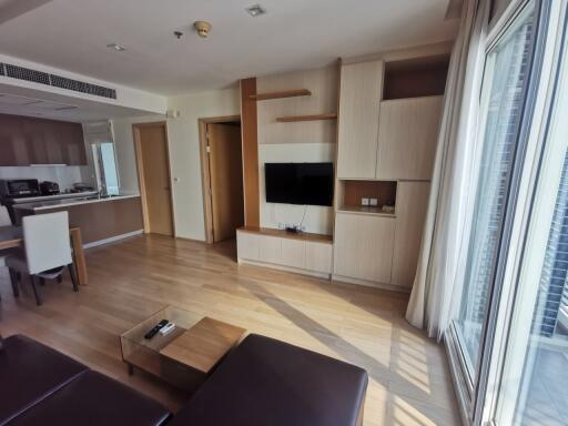 Modern living room with TV and wooden furniture, adjacent to an open kitchen.