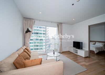 Living room with a beige sofa, wall-mounted TV, a window with a city view, and an adjacent bedroom