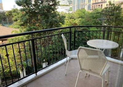 A cozy balcony with a small table and two chairs, overlooking a green area and city buildings.